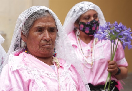 Se muestra en la imagen dos mujeres indigenas, con un velo de tela blanca, sosteniendo flores moradas. Una de ella es Ifigenia González; maestra, promotora cultural y artesana nahua, Ifigenia rompió silencios con cada danza y cada puntada.Durante décadas, ha preservado tradiciones desde lo femenino, resignificando el papel de las mujeres indígenas como creadoras de cultura viva.