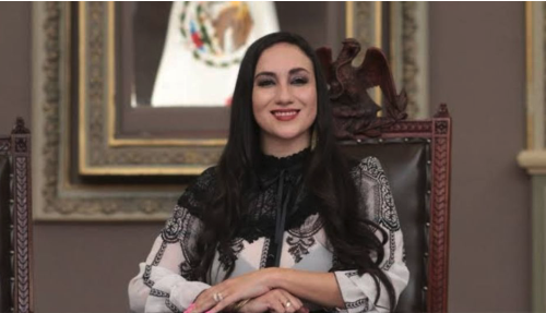 Retrato de una mujer joven de tez clara y cabello negro largo y ondulado, sonriendo con los labios pintados de rosa. Viste una blusa elegante de encaje negro y tela estampada en blanco y negro. Está sentada en un sillón de cuero con detalles tallados, con el Escudo Nacional Mexicano en un cuadro al fondo.