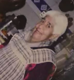 Mujer de edad avanzada, con cabello blanco, sonriendo cálidamente. Está envuelta en un mandil rayado, con una prenda oscura y estampada debajo. El fondo muestra un patrón de azulejos blancos y azules lo que aparenta ser una cocina.